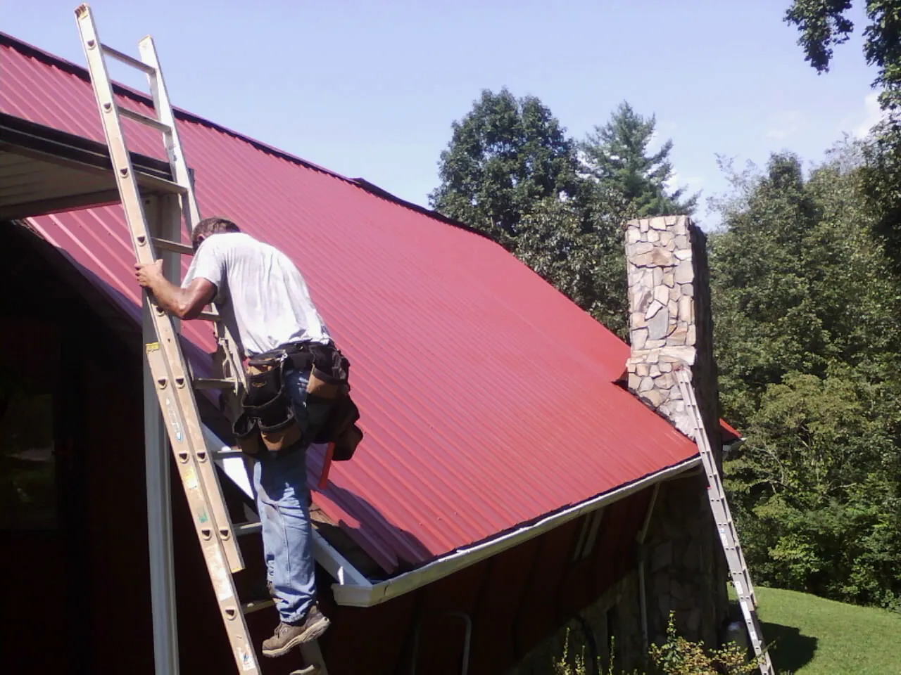 Roofer on a ladder beside a red metal roof and stone chimney — Soffit & Fascia Repair by Roofing Brothers in Overland Park